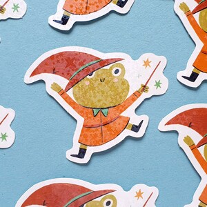 May include: A collection of whimsical stickers featuring a frog dressed as a wizard. The frog is wearing an orange coat and a red hat with star patterns, holding a wand. The stickers are on a light blue background.