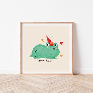 Puede incluir: Una impresión artística enmarcada con una ilustración de dibujos animados de una rana verde con un gorro de fiesta. La rana está adornada con un corazón rojo y estrellas, con la frase "nice butt" escrita debajo. La obra de arte está sobre un fondo crema.