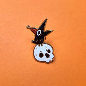 May include: A metal enamel pin featuring a black bird wearing a red party hat with a small star, perched atop a white human skull. The skull has black eye sockets and a nose cavity, with gold-toned metal outlines defining the entire design.