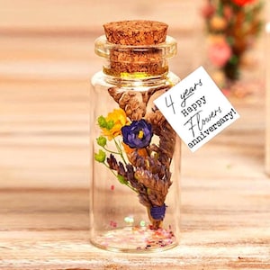 Puede incluir: Una pequeña botella de vidrio transparente con tapón de corcho, llena de flores secas y una etiqueta que dice "4 years Happy Flowers anniversary!". Las flores son amarillas, moradas y marrones, con confeti en el fondo.
