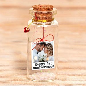 Puede incluir: Una botella de vidrio transparente con un tapón de corcho, que contiene pequeñas partículas blancas y una foto de una pareja. La foto está sujeta con un hilo rojo y tiene el texto "Happy 1st anniversary!". Un pequeño dije de corazón rojo cuelga de la botella.