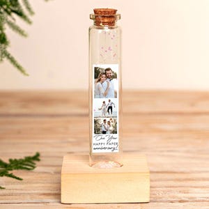 Puede incluir: Una botella de vidrio transparente con un tapón de corcho, que contiene pequeñas decoraciones rosas y una tira de fotos. La tira de fotos muestra tres imágenes de una pareja y el texto "One Year Happy Paper Anniversary!" La botella se encuentra sobre un pequeño bloque de madera.