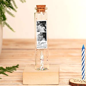 Puede incluir: Una botella de vidrio transparente con tapón de corcho, que contiene una tira de fotos y confeti, se encuentra sobre un bloque de madera. La tira de fotos dice "16 years HAPPY WAX anniversary!". Una vela a rayas azules y blancas está sobre una rodaja de madera.