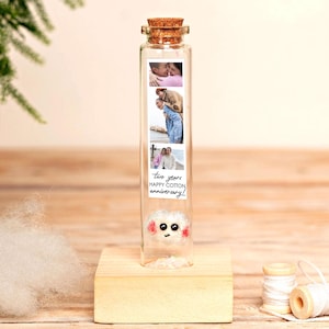 May include: A clear glass test tube with a cork stopper, mounted on a square wooden base. Inside is a vertical strip of three photographs and the text "two years HAPPY COTTON anniversary!" above a small, white, fluffy creature with pink cheeks.