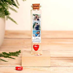 Puede incluir: Una botella de vidrio transparente con un tapón de corcho, que contiene un corazón rojo y fotos. La botella está sobre un bloque de madera. Las fotos muestran parejas. El texto en la botella dice "9 years happy pottery anniversary!"