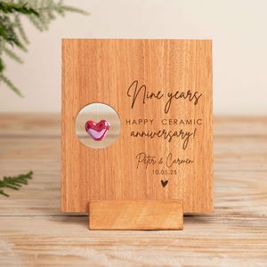 Puede incluir: Placa de aniversario de madera con las palabras "Nine years HAPPY CERAMIC anniversary!" y los nombres "Peter & Carmen" y una fecha. Un pequeño corazón rojo cuelga en una ventana circular.