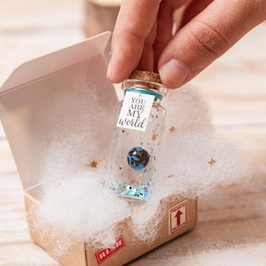 Puede incluir: Una pequeña botella de vidrio con tapón de corcho, que contiene un planeta en miniatura y purpurina, se sostiene sobre una caja de cartón abierta. La botella tiene una etiqueta que dice "YOU ARE MY world". La caja está llena de pelusa blanca.