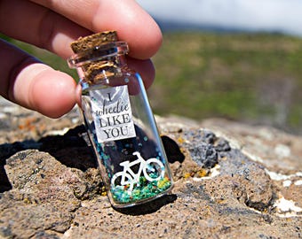 May include: A small glass bottle with a cork stopper, filled with colorful confetti and a bicycle silhouette. The bottle has a label that reads "I wheelie LIKE YOU". The bottle is held against a blurred natural background.