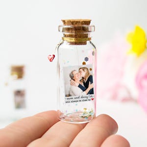 Puede incluir: Una pequeña botella de vidrio transparente con tapón de corcho, que contiene una foto de una madre y su hija. La botella está decorada con confeti y un dije de corazón rojo. El texto en la foto dice "mamá e hija siempre estarán cerca."