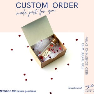 May include: A clear glass bottle with a cork stopper inside a small cardboard box. The box is open, revealing white filler and red heart-shaped confetti. Text on the box reads "CUSTOM ORDER made just for you". The bottle has a tag with text.