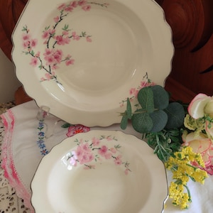 May include: Two off-white ceramic serving bowls with scalloped edges. Each bowl features a delicate pink cherry blossom design with black branches. The rims have a silver trim. The bowls are on a white cloth.