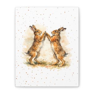 May include: Watercolor painting of two brown hares standing on their hind legs, touching paws. The hares are depicted with detailed fur and are set against a soft, neutral background with subtle watercolor washes and scattered speckles.