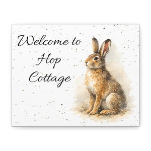 May include: A canvas print featuring a watercolor illustration of a brown rabbit. The text "Welcome to Hop Cottage" is written in elegant script. The background is white with subtle speckled accents.