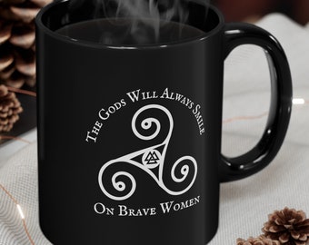 May include: Black coffee mug with a white design featuring a triquetra symbol and the text "The Gods Will Always Smile On Brave Women".