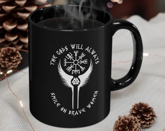 May include: Black ceramic mug with a white graphic featuring a Viking compass, wings, and the text "THE GODS WILL ALWAYS SMILE ON BRAVE WOMEN". The mug is filled with a dark liquid, possibly coffee or tea.