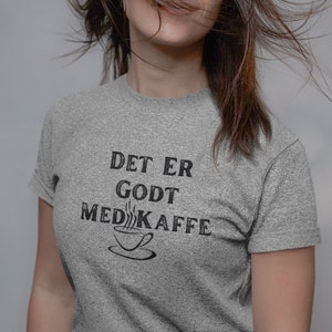 May include: A woman wearing a grey t-shirt with the text "Det er godt med kaffe" and a coffee cup graphic.