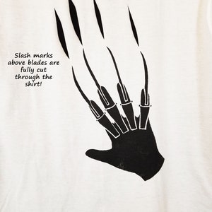 May include: White t-shirt featuring a black graphic of a hand with long, sharp claws. The text "Slash marks above blades are fully cut through the shirt!" is printed below the claws. The design is a horror-themed graphic.