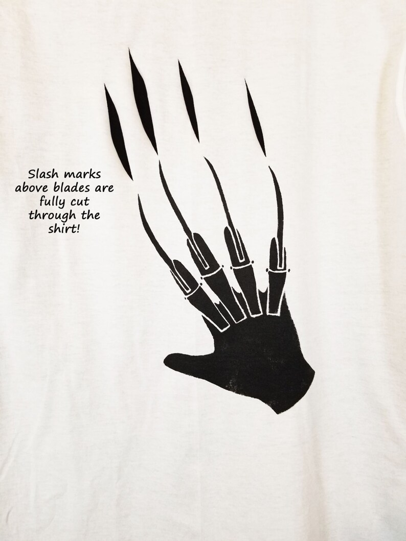 May include: White t-shirt featuring a black graphic of a hand with long, sharp claws. The text "Slash marks above blades are fully cut through the shirt!" is printed below the claws. The design is a horror-themed graphic.