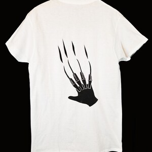 May include: White t-shirt featuring a black graphic of a hand with long, sharp claws. The clawed hand design is printed on the back of the shirt. The shirt is made of a soft, lightweight material.
