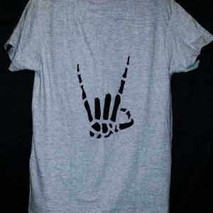 Może przedstawiać: Szary T-shirt z czarnym szkieletowym ręką wykonującą gest rock and roll.
