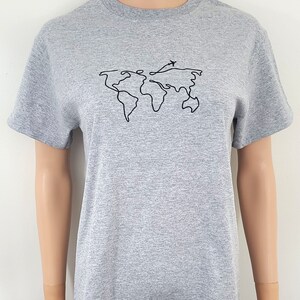 K&ouml;nnte beinhalten: Ein graues T-Shirt mit einer schwarzen Linienzeichnung der Welt mit einem kleinen Flugzeug, das dar&uuml;ber fliegt.