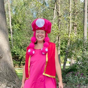 May include: A bright pink sleeveless dress with a gold trim at the hem, paired with a red vest featuring gold edges. The outfit includes a pink mushroom-shaped hat with white spots and two long, dangling pink braids with white polka dots.