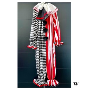 May include: A full-body clown costume with a black and white diamond pattern on one side and red and white stripes on the other. The costume has a white ruffled collar, black pom-poms, and red trim. The sleeves and pant legs are flared.