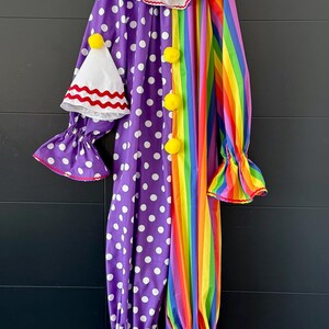 May include: A colorful clown costume with a white ruffled collar and a matching hat. The costume features a purple polka-dot pattern on one side and rainbow stripes on the other. Yellow pom-poms and red trim add detail.