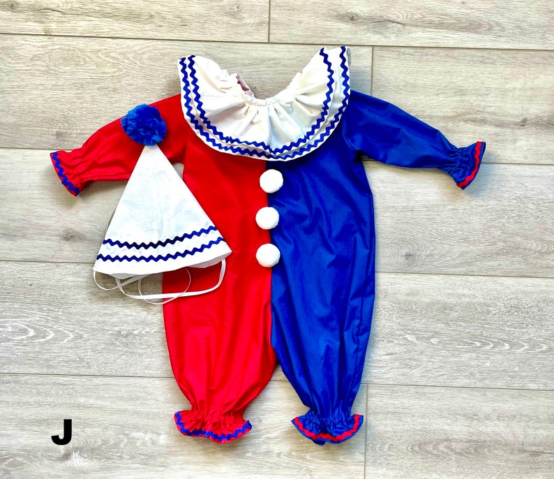 May include: A red and blue clown costume with a matching hat. The one-piece outfit has a white ruffled collar and three white pom-pom buttons. The hat is white with a blue zig-zag trim and a blue pom-pom.