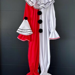 May include: A red and white clown costume with a ruffled collar and cuffs, black pom-pom buttons, and a matching hat. The costume is hanging on a white hanger against a dark background. The costume is made of soft fabric.