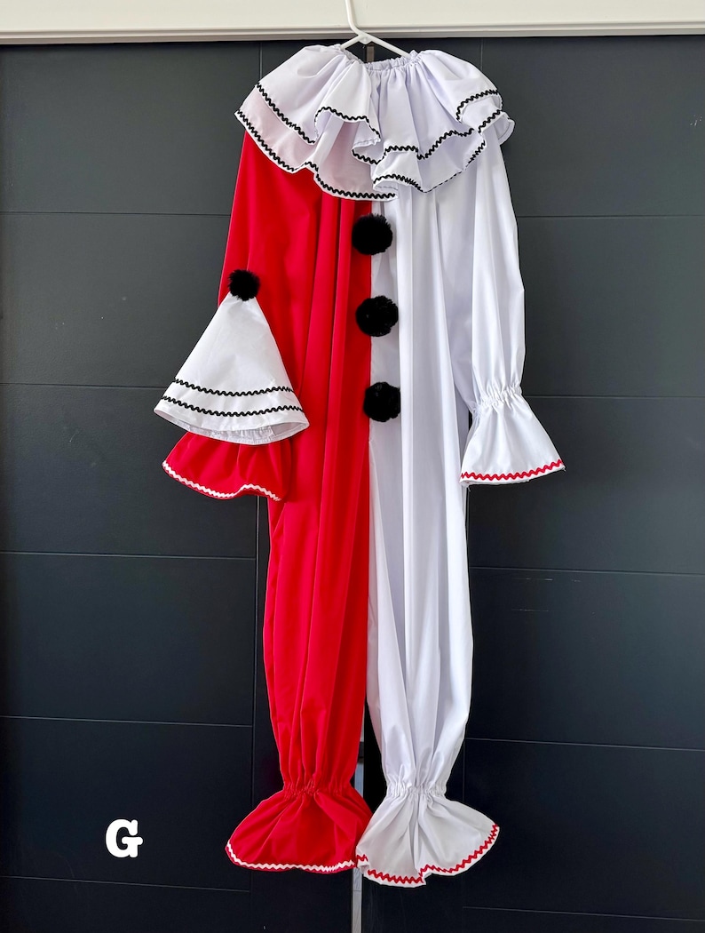 May include: A red and white clown costume with a ruffled collar and cuffs, black pom-pom buttons, and a matching hat. The costume is hanging on a white hanger against a dark background. The costume is made of soft fabric.