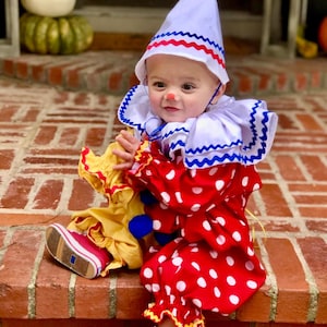 May include: A child's clown costume featuring a white hat with red and blue trim, a ruffled collar, and a red and white polka-dot outfit. The costume includes red shoes and a red nose, perfect for Halloween or a costume party.