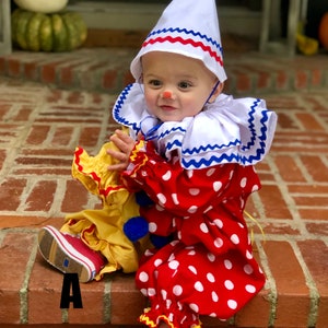 K&ouml;nnte beinhalten: Ein Baby, das als Clown verkleidet ist, tr&auml;gt einen rot-wei&szlig; gepunkteten Overall mit wei&szlig;em Kragen und blauen Besatz, einen wei&szlig;en Hut mit rotem und blauem Besatz und rote Schuhe. Das Baby sitzt auf einer Ziegelstufe.