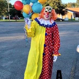 May include: A person in a colorful clown costume holding a bunch of balloons. The costume is split down the middle, with one side yellow and the other red with white polka dots. The clown has orange hair and a painted face.