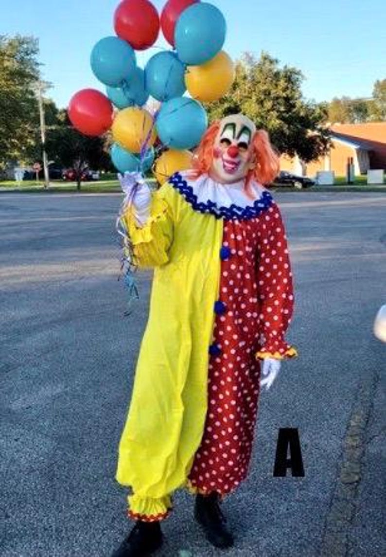 May include: A person in a colorful clown costume holding a bunch of balloons. The costume is split down the middle, with one side yellow and the other red with white polka dots. The clown has orange hair and a painted face.
