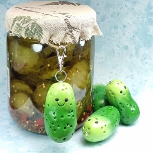 May include: A glass jar filled with sliced pickles, topped with a beige fabric lid cover. A green pickle-shaped charm with a smiling face and a silver lobster clasp hangs from the jar. Two additional green pickle charms sit nearby.
