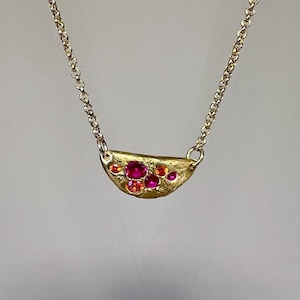 May include: A gold-toned necklace featuring a crescent-shaped pendant adorned with vibrant red and orange gemstones. The pendant is suspended from a delicate chain, creating an elegant and eye-catching piece of jewelry.