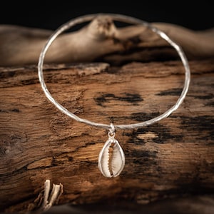 Op de afbeelding: Een zilverkleurige choker ketting met een getextureerde band en een schelpenhanger. De hanger is een wit en bruin schelpje in een zilveren setting. De ketting wordt getoond op een stuk verweerd hout.