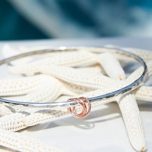 Könnte beinhalten: Ein silbernes Armreif-Armband mit gehämmerter Oberfläche und einem roségoldenen Akzent. Das Armband liegt auf einem weißen Seestern.