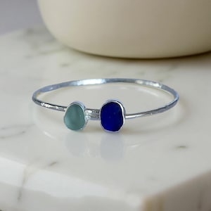 Può includere: Un bracciale rigido in argento con due pietre di vetro marino. Una è verde acqua chiaro, l'altra blu scuro. Il bracciale ha una texture martellata e un design semplice. Circonferenza di circa 18 cm.