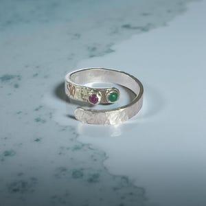 May include: A silver ring with a hammered finish, set with a red and a green gemstone. The ring has an open design, with the gemstones set close together on the band.