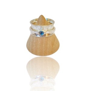 Peut inclure: Une bague en argent avec une pierre précieuse bleue sertie au centre, empilée sur deux autres bandes en argent. La bague est présentée sur un porte-bague en bois.