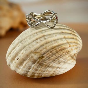 May include: A silver-toned ring with an intricate, interwoven design, featuring small red and green gemstones. The ring rests on a large, textured seashell, with a warm, neutral background.