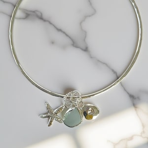 Puede incluir: Un collar plateado con un diseño circular. El collar presenta una estrella de mar, una piedra azul claro y un pequeño dije en forma de corazón. El collar se exhibe sobre una superficie de mármol blanco.