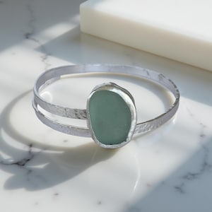 May include: A silver bangle bracelet featuring a seafoam green, oval-shaped stone. The bracelet has a hammered texture and a double-band design. The stone is set in a silver bezel. The bracelet is a piece of jewellery.
