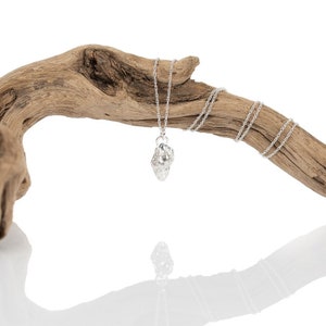 May include: A silver necklace with a textured, organic-shaped pendant. The necklace is displayed on a piece of driftwood.