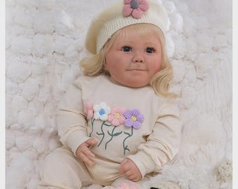 May include: A realistic baby doll with blonde hair wearing a cream-colored long-sleeve shirt and pants set, a matching beret with a pink flower, and cream socks with brown accents. The shirt features embroidered flowers and the text The Newborn Nebula Nursery.