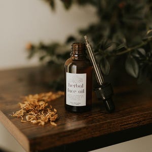 May include: A brown glass bottle of herbal face oil with a dropper, set on a dark wooden surface. The label reads "herbal face oil" with additional text. Dried flower petals are scattered nearby. The bottle contains 1 fl oz / 29ml.