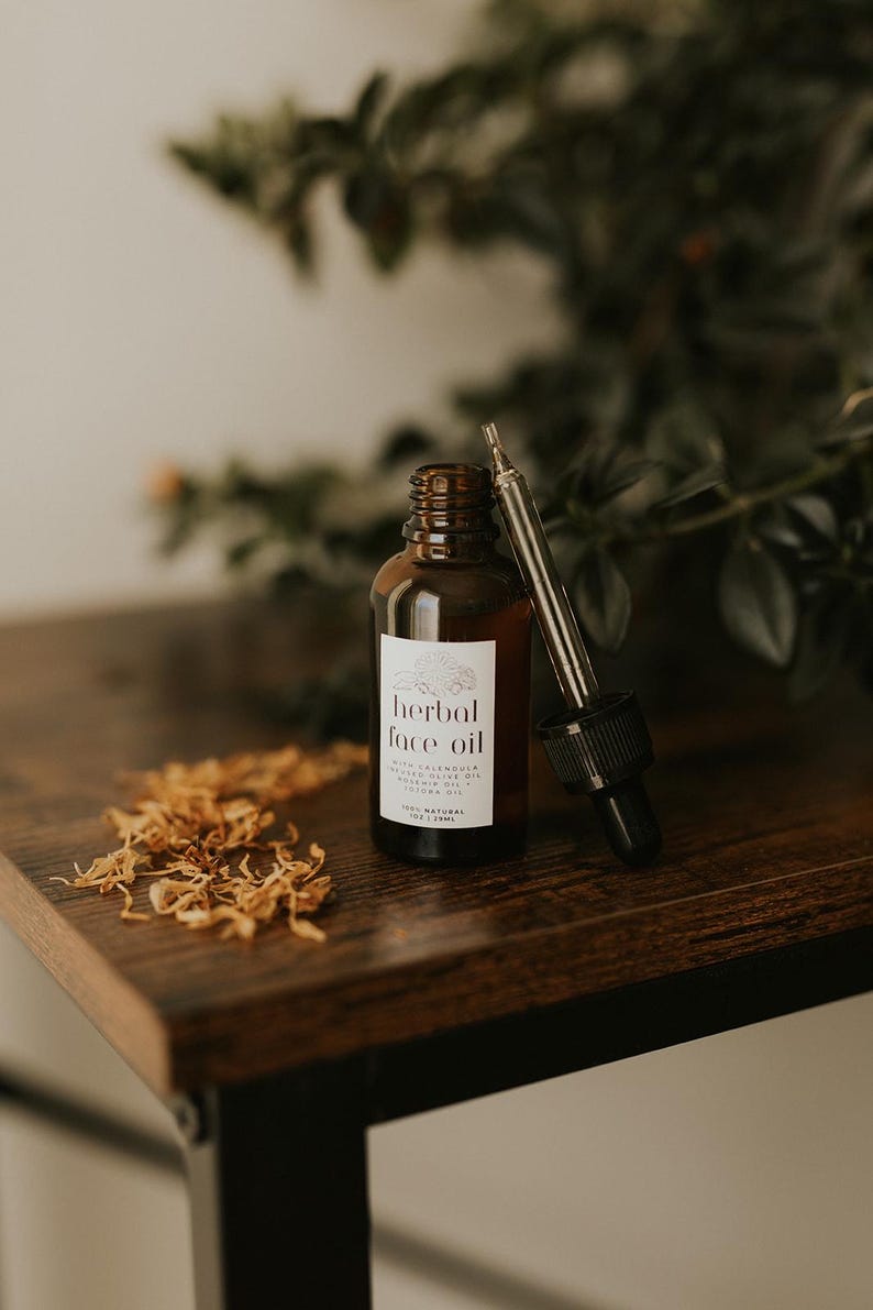 May include: A brown glass bottle of herbal face oil with a dropper, set on a dark wooden surface. The label reads "herbal face oil" with additional text. Dried flower petals are scattered nearby. The bottle contains 1 fl oz / 29ml.