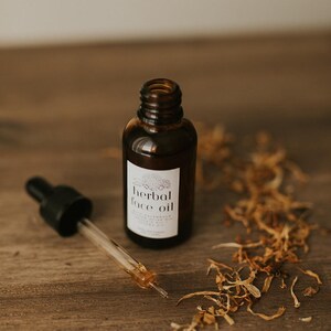 May include: A brown glass bottle of herbal face oil with a dropper, set on a wooden surface. The label reads "herbal face oil". Dried plant matter is scattered around the bottle and dropper.